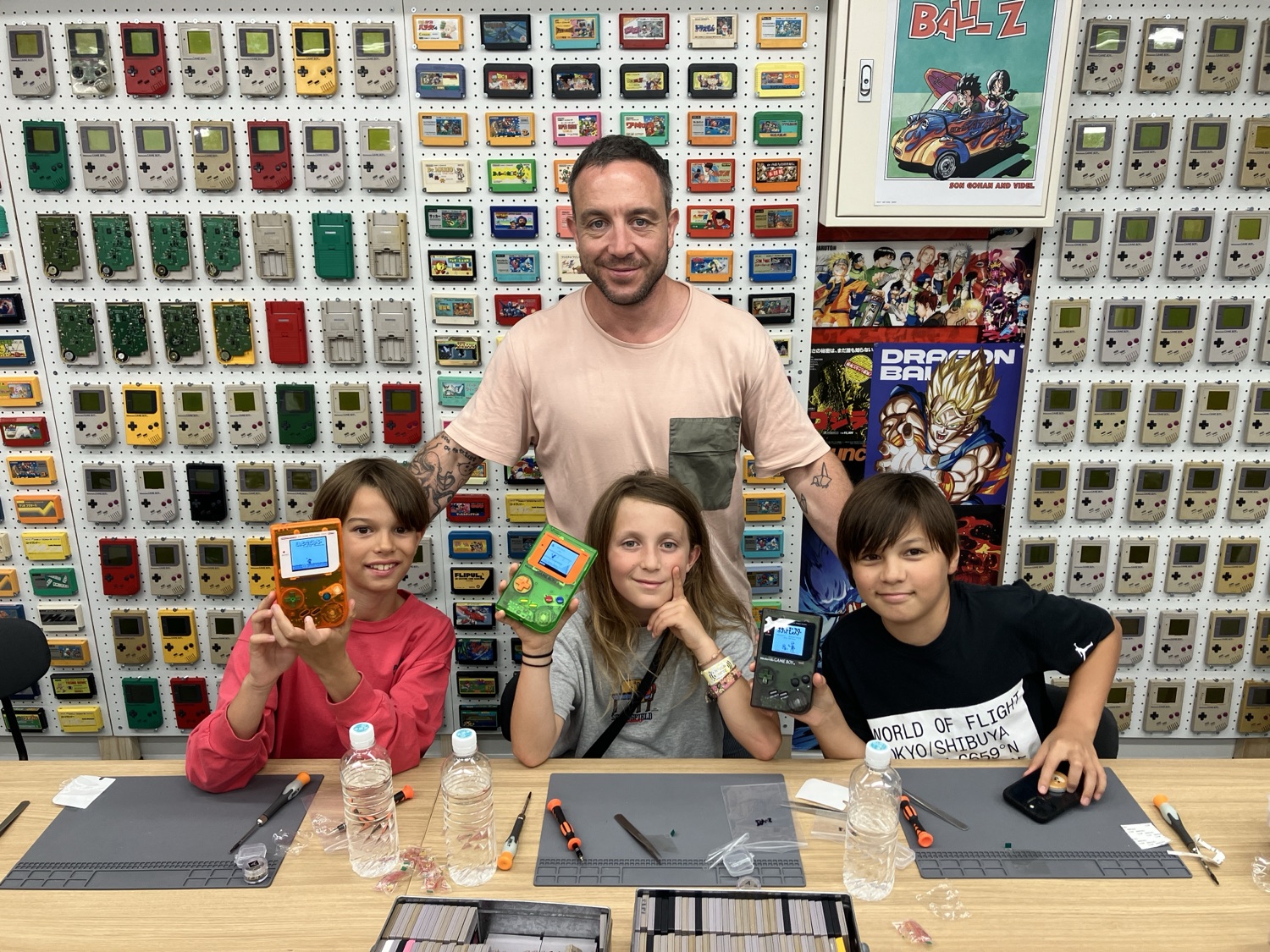 A dad and three kids proudly holding up the Game Boys they just built, in front of the cartridge wall