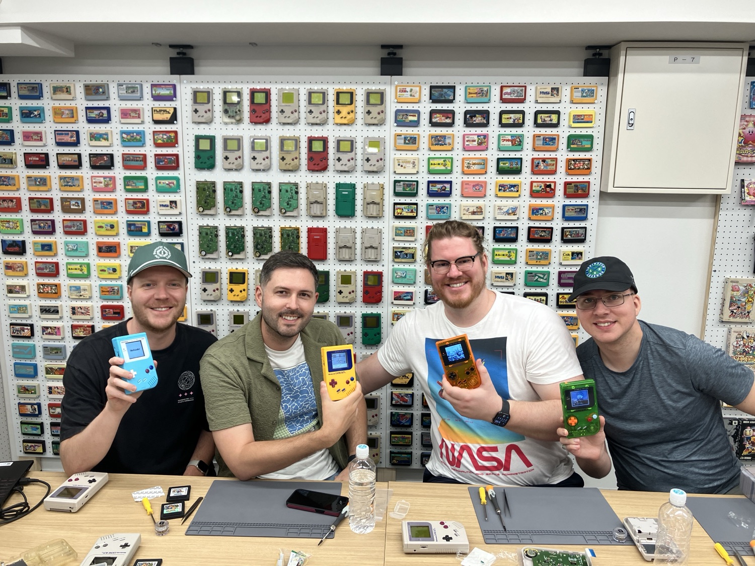 Four friends holding up four colorful finished Game Boys in front of the cartridge wall