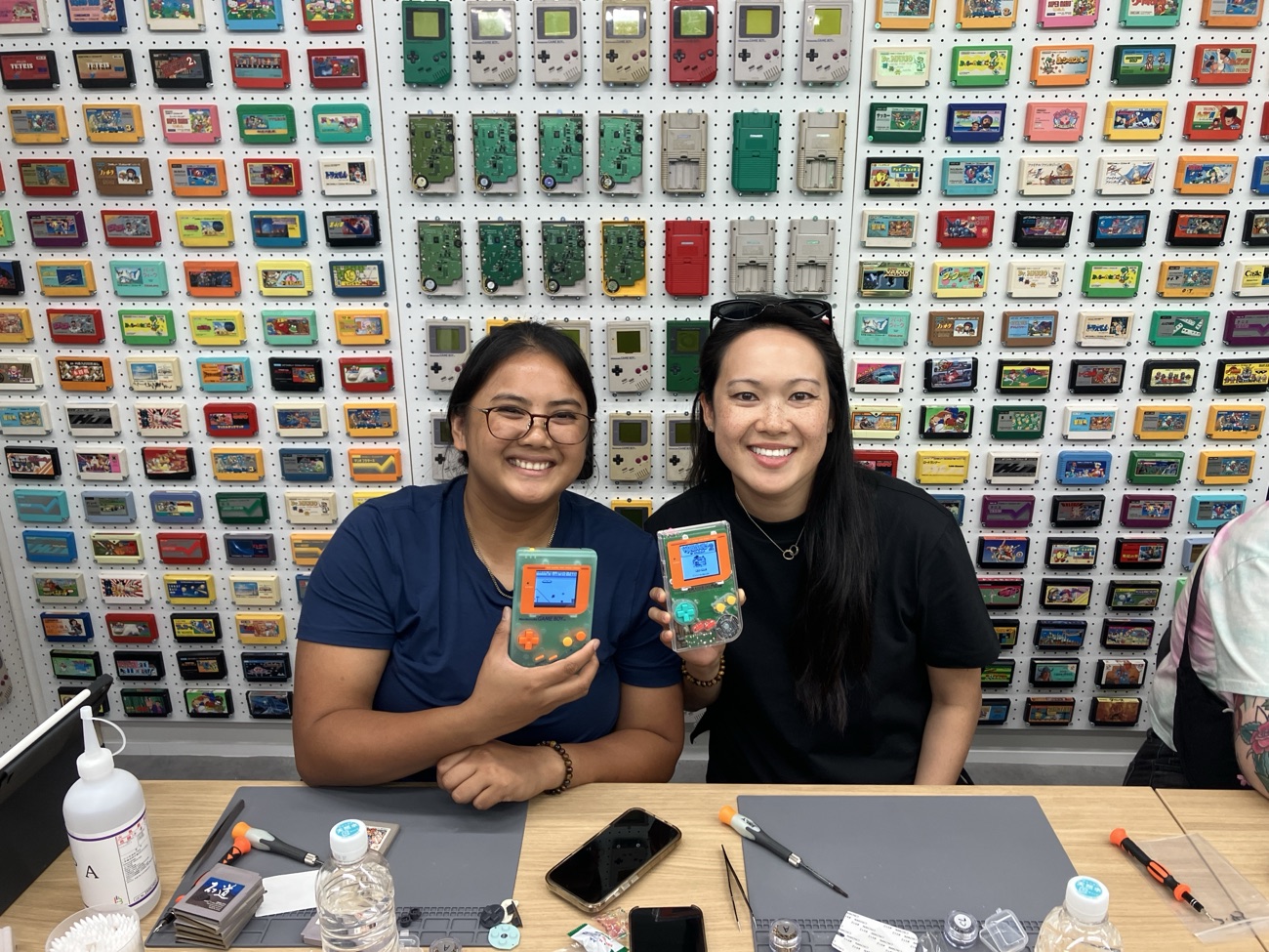 Two friends smiling with their finished clear-orange Game Boys in front of the cartridge wall